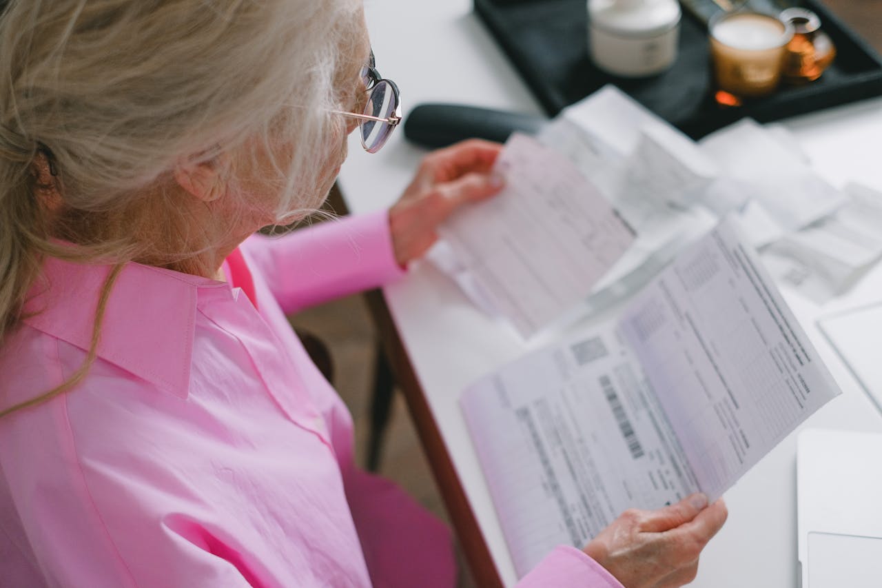 An elderly woman in glasses holds and reads important papers at a table indoors.
