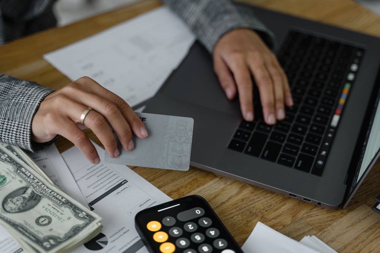 Close-up of a person using a credit card and laptop in a financial setting with cash and calculator.
